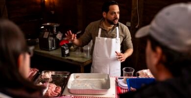 El Instructor de carnicería Marcos somana hablando con un grupo de personas alrededor de una mesa en un taller de carnicería rústico. Está explicando el proceso mientras manipula varios tipos de carne en un entorno iluminado de forma cálida y natural.