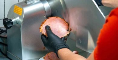 Marcos somana utilizando una máquina cortadora de fiambre para rebanar carne curada, con guantes negros para mantener la higiene. Las lonchas de carne caen ordenadamente en un plato sobre una superficie de azulejos azules. En el fondo se observan embutidos colgando, creando un ambiente de charcutería artesanal.