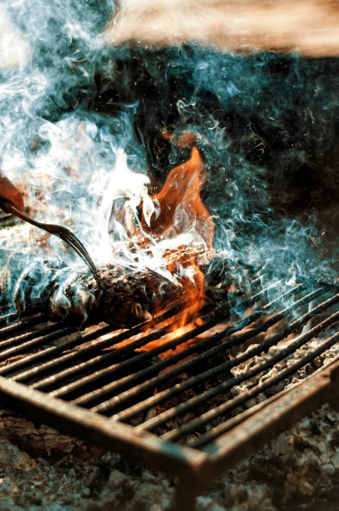 Parrilla encendida con una pieza de carne que se cocina sobre llamas y humo denso. Una mano, apenas visible, sostiene un tenedor, dándole la vuelta a la carne mientras el fuego resplandece, creando una atmósfera intensa y rústica.