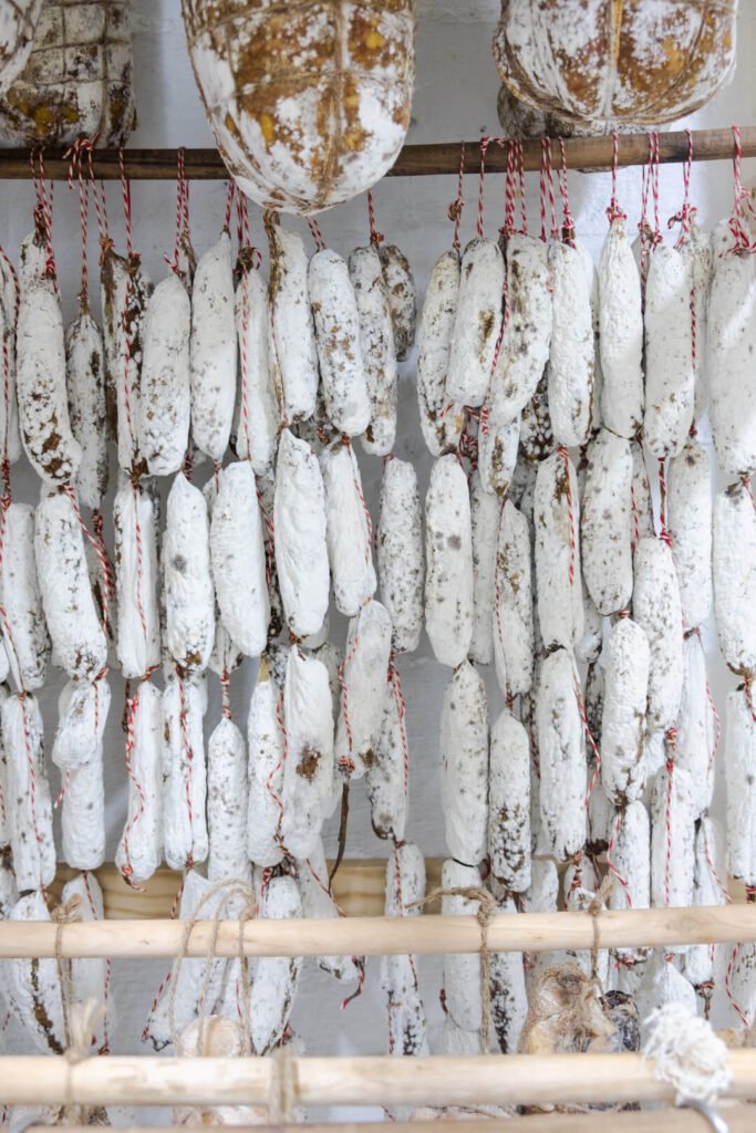 Salames blancos cubiertos de moho noble, colgando en filas perfectamente alineadas en una sala de curado. Estas piezas muestran un proceso artesanal de maduración tradicional que garantiza un sabor profundo y una textura óptima. Elaborados con dedicación por Marcos Somana, creador de La Fiambrería y maestro charcutero.