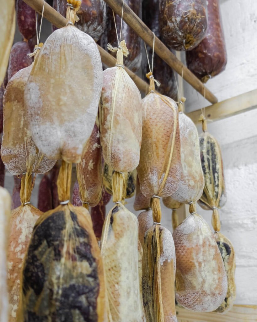 Piezas de prosciutto de pato cuidadosamente colgadas con cuerdas en un espacio diseñado para el curado. Las piezas tienen una textura exterior que refleja un proceso de maduración preciso, resaltando sabores complejos y exclusivos. Este producto gourmet es una creación de Marcos Somana, reconocido maestro charcutero y fundador de La Fiambrería.